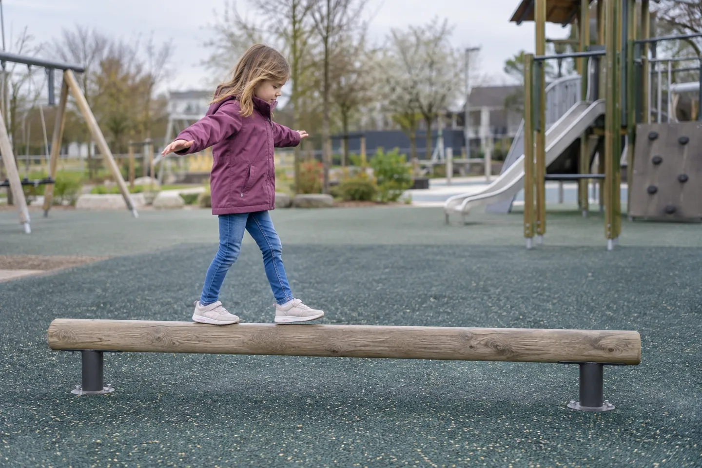 A child crosses a fixed timber balance beam bolted over rubber safety surfacing at a public playground, where standards cap the beam's height for each age band instead of regulating width.