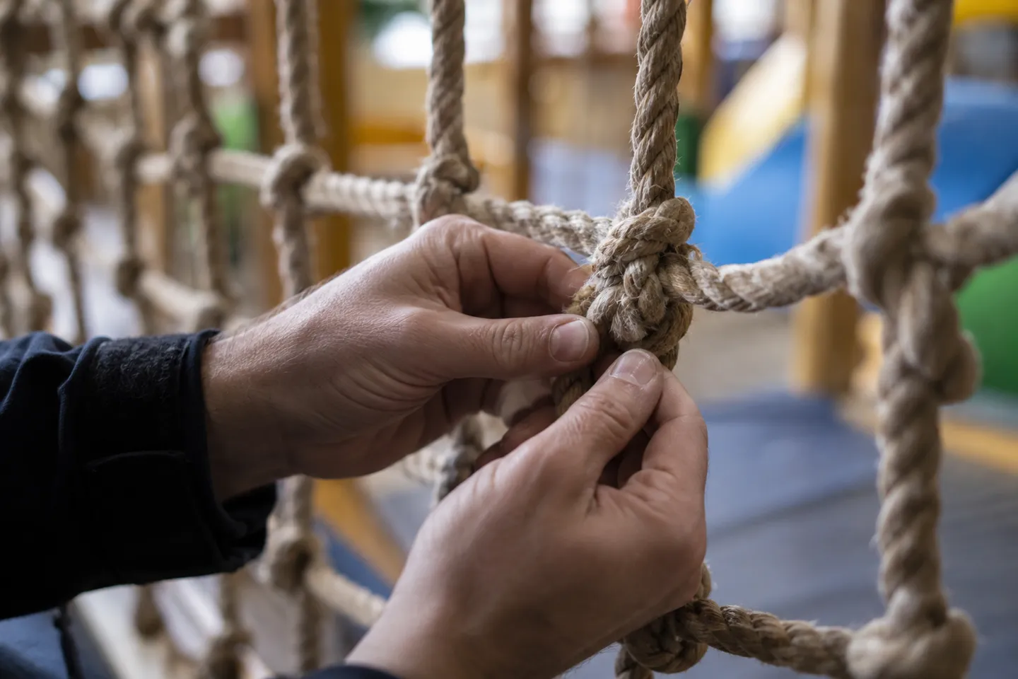 Close-up of hands checking the knots of a climbing rope net for fraying during a routine indoor playground inspection